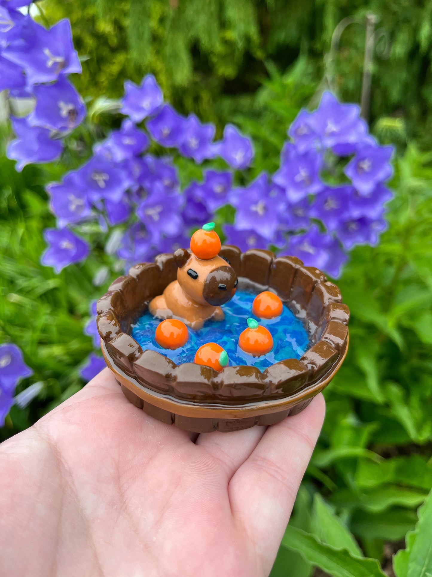 A hand holding a small ceramic trinket dish shaped like a capybara, with orange and blue colors and adorned with fake oranges.